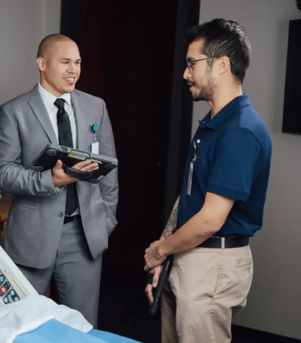 Two men chatting, one in a suit holding a tablet and the other casually dressed, in an office setting.