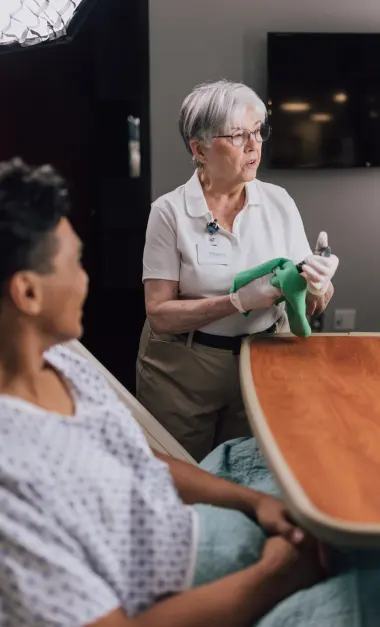 An elderly woman in a hospital gown sits at a table while a nurse holds a spirometer, assisting with a breathing exercise.